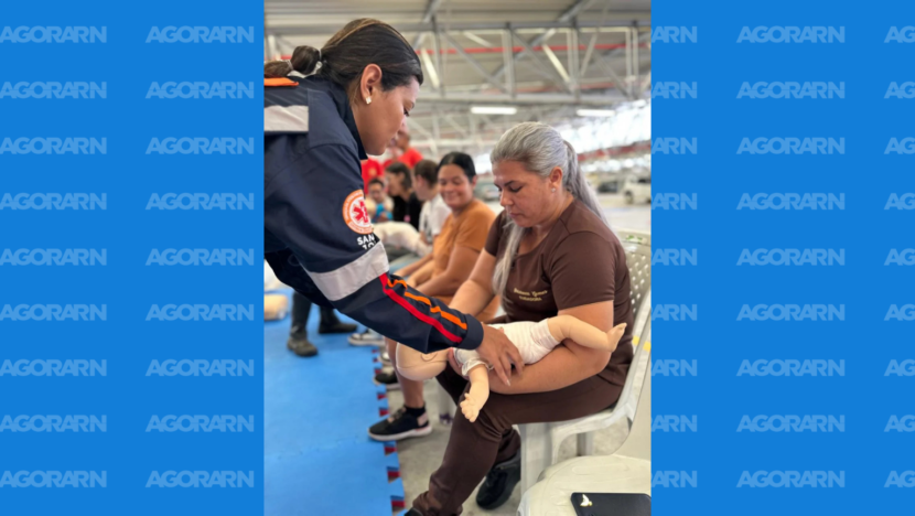 Profissional uniformizada do SAMU demonstra procedimentos de primeiros socorros em manequim infantil, enquanto uma mulher acompanha a atividade sentada em uma cadeira