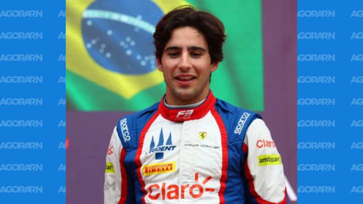 Piloto de automobilismo com macacão branco, vermelho e azul, com logotipos da Ferrari, Claro e patrocinadores, diante de uma bandeira do Brasil