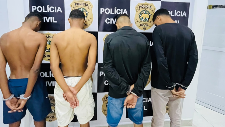Quatro homens fotografados de costas, com as mãos algemadas, diante de painel com identificação da Polícia Civil do Rio Grande do Norte