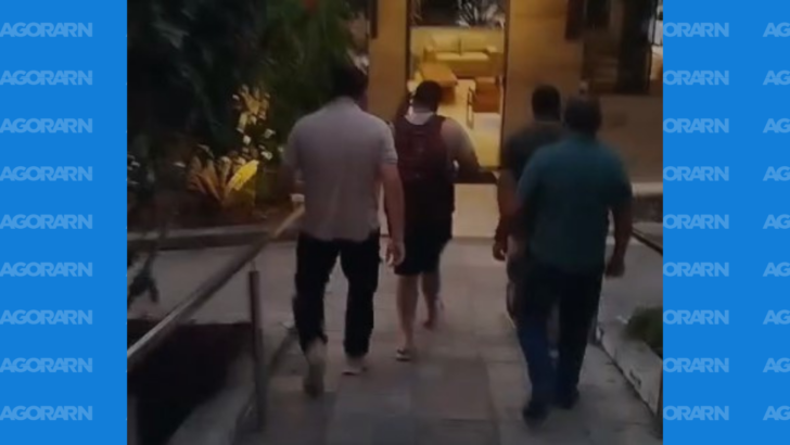 Três homens caminham por uma passagem externa em direção à entrada de um prédio, com uma pessoa de camiseta vermelha ao centro