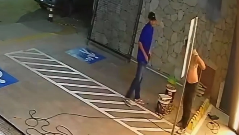 Imagem de câmera de segurança mostra um homem de camisa azul próximo a outro homem junto à entrada de um imóvel, em uma área com vagas de estacionamento demarcadas