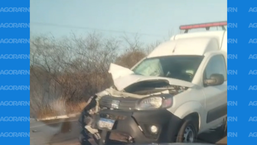 Ambulância branca parada em uma rodovia, com capô amassado, para-choque dianteiro danificado e parte do motor exposta