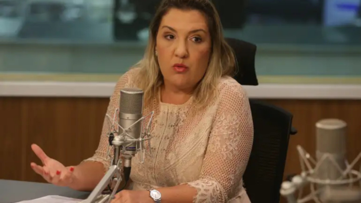 Mulher de cabelos loiros, vestindo blusa clara, fala diante de um microfone em um estúdio de rádio