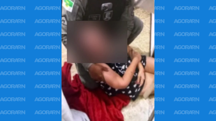 Mulher sentada no chão, com o rosto desfocado, recebe assistência de uma pessoa uniformizada em ambiente interno. Um tecido vermelho aparece próximo a ela