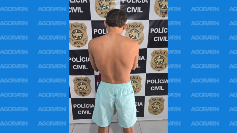 Homem sem camisa, vestindo bermuda azul-clara, aparece de costas diante de um painel com o nome e o brasão da Polícia Civil do Rio Grande do Norte