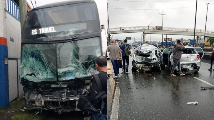 Ônibus de dois andares com a parte frontal destruída e para-brisa estilhaçado aparece ao lado de dois carros com danos graves após acidente em rodovia. Pessoas observam os veículos, enquanto uma passarela aparece ao fundo.