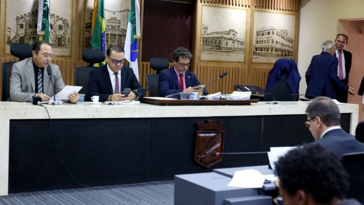 Três vereadores sentados à mesa de reunião da Câmara de Natal, com documentos, celulares e microfones