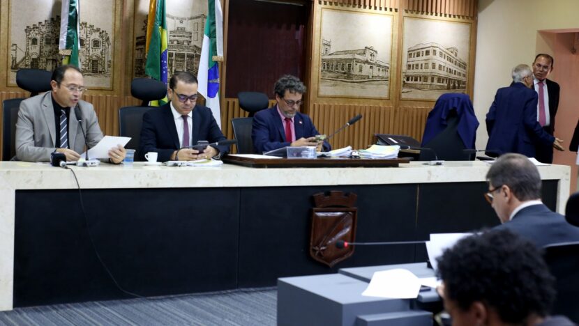 Três vereadores sentados à mesa de reunião da Câmara de Natal, com documentos, celulares e microfones