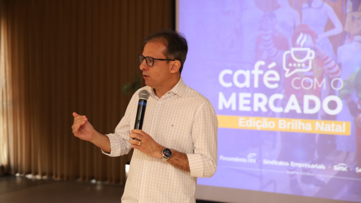 Homem de óculos segura microfone diante de uma tela com a identificação Café com o Mercado, edição Brilha Natal