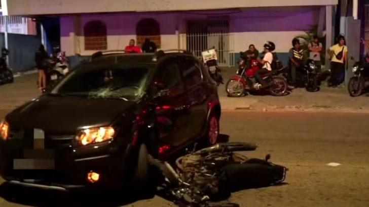 Carro preto com para-brisa danificado e motocicleta caída junto à parte dianteira, à noite, com pessoas na calçada ao fundo