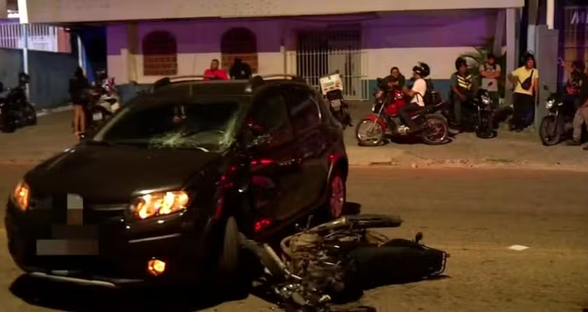 Carro preto com para-brisa danificado e motocicleta caída junto à parte dianteira, à noite, com pessoas na calçada ao fundo