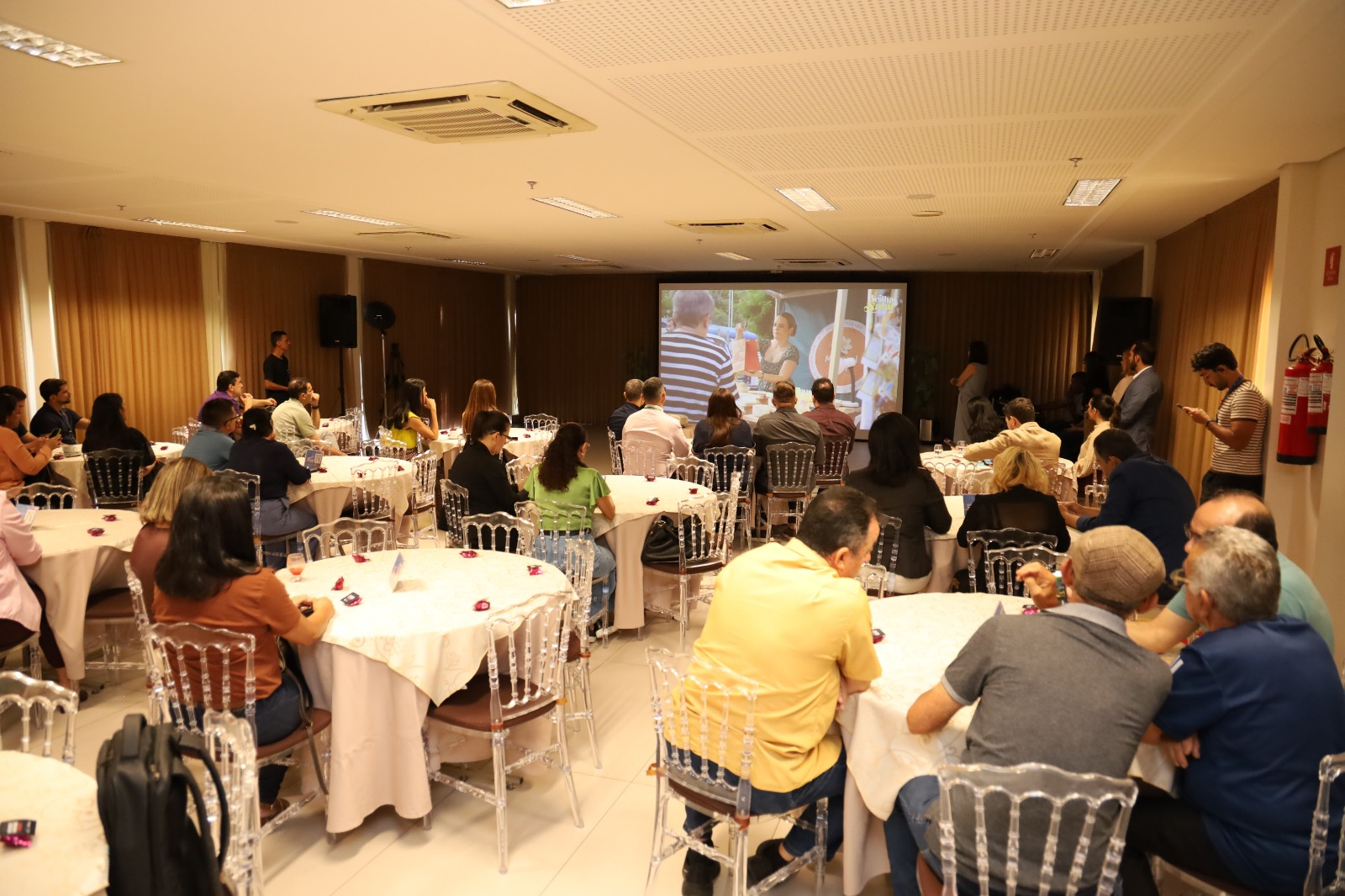 Participantes sentados em mesas acompanham uma apresentação em telão durante o Café com o Mercado, no Sesc Rio Branco