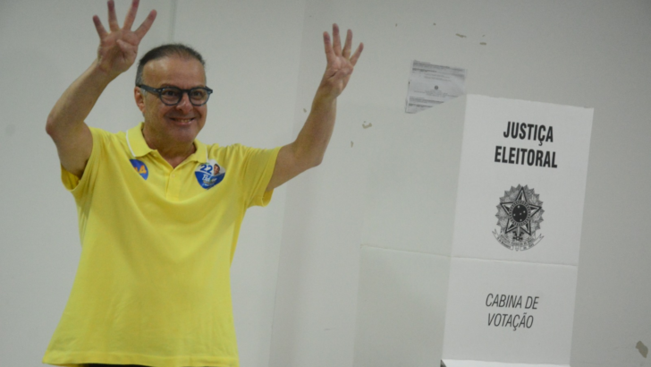 Paulinho Freire, de camisa amarela e óculos, levanta as mãos ao lado de uma cabina de votação