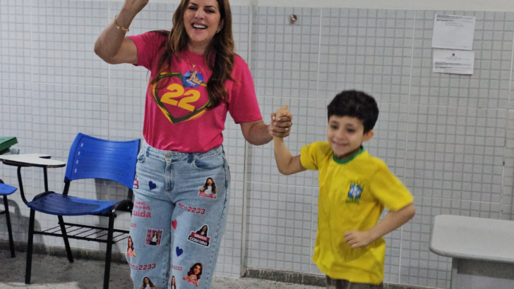 Nina Souza, de camiseta rosa e calça jeans, faz sinal de vitória e segura a mão de uma criança em uma sala de aula