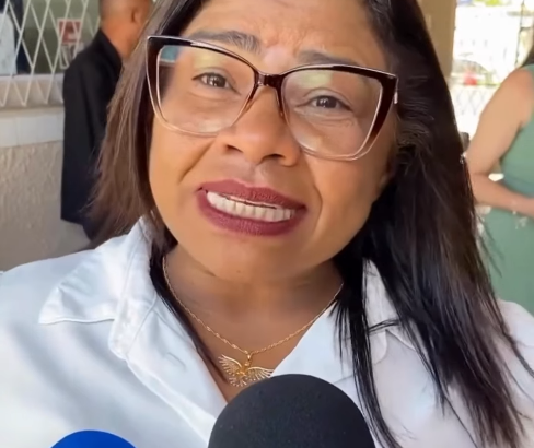 Nilda Cruz, de óculos e camisa branca com adesivos de campanha, fala diante de um microfone
