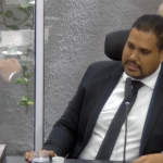 Michael Diniz, de terno escuro e gravata, sentado e falando ao microfone na Câmara de Parnamirim