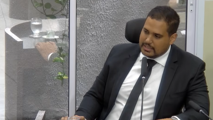Michael Diniz, de terno escuro e gravata, sentado e falando ao microfone na Câmara de Parnamirim
