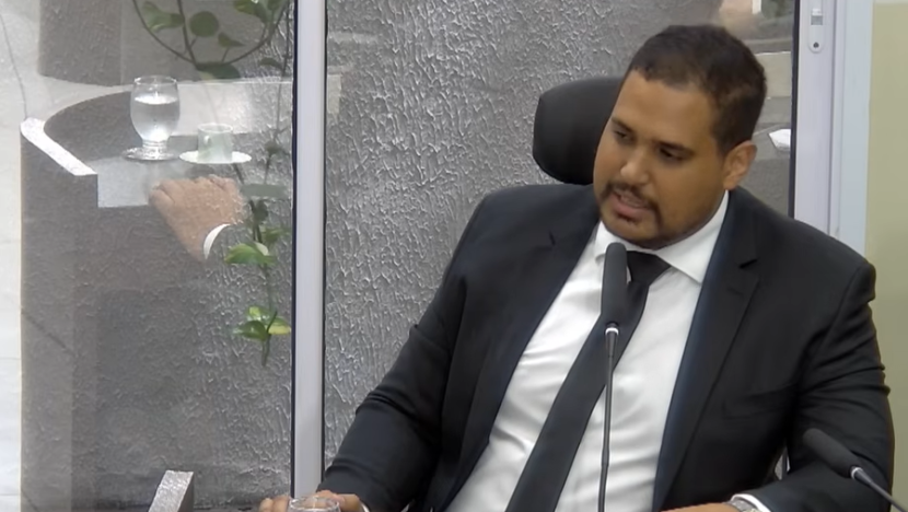 Michael Diniz, de terno escuro e gravata, sentado e falando ao microfone na Câmara de Parnamirim