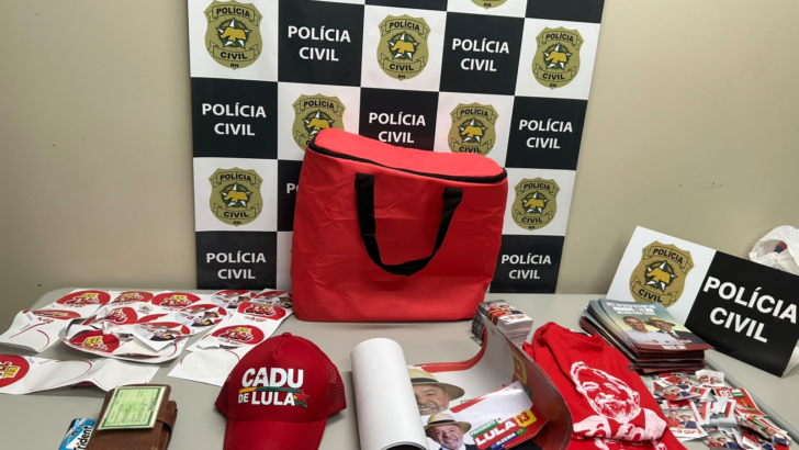 Boné, camisetas, adesivos e panfletos de campanha, bolsa vermelha e carteira sobre mesa diante de painel da Polícia Civil