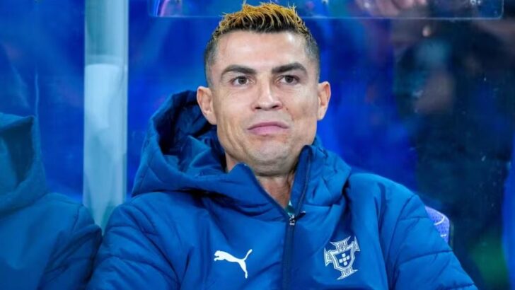 Cristiano Ronaldo, de agasalho azul acolchoado com o escudo da seleção portuguesa, sentado no banco de reservas com expressão fechada