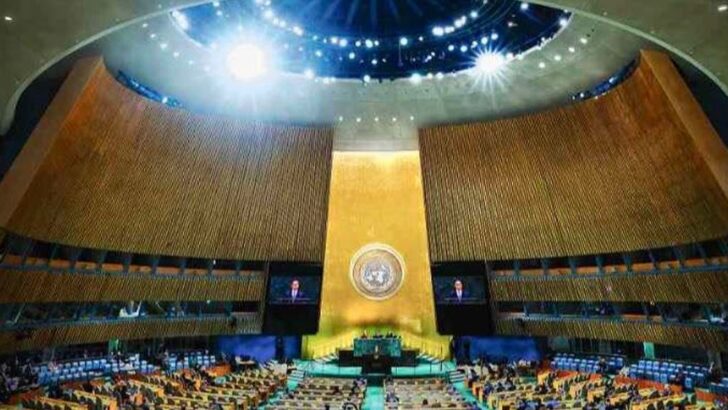 Vista do alto do plenário da Assembleia Geral das Nações Unidas, com a cúpula iluminada, a parede dourada e o emblema da ONU atrás da tribuna, e as bancadas ocupadas