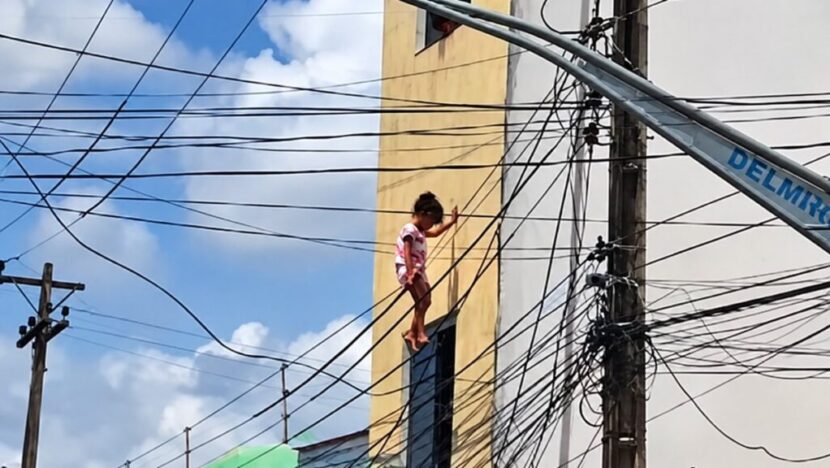 Criança aparece suspensa entre fios de uma rede aérea, próxima à fachada de um prédio de dois andares, em rua com postes e cabos elétricos