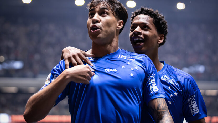 Dois jogadores do Cruzeiro comemoram abraçados; o atleta em primeiro plano puxa a camisa azul na altura do peito.