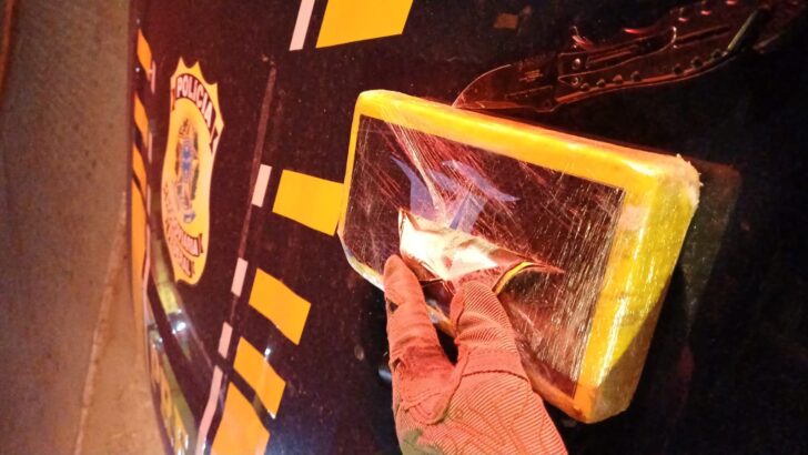 Mão com luva segura um pacote retangular envolvido em plástico transparente e fita amarela diante de uma viatura da Polícia Rodoviária Federal, durante a noite