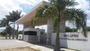 Fachada da Escola Agrícola de Jundiaí, com entrada coberta, guarita, palmeiras e identificação da UFRN na parede externa