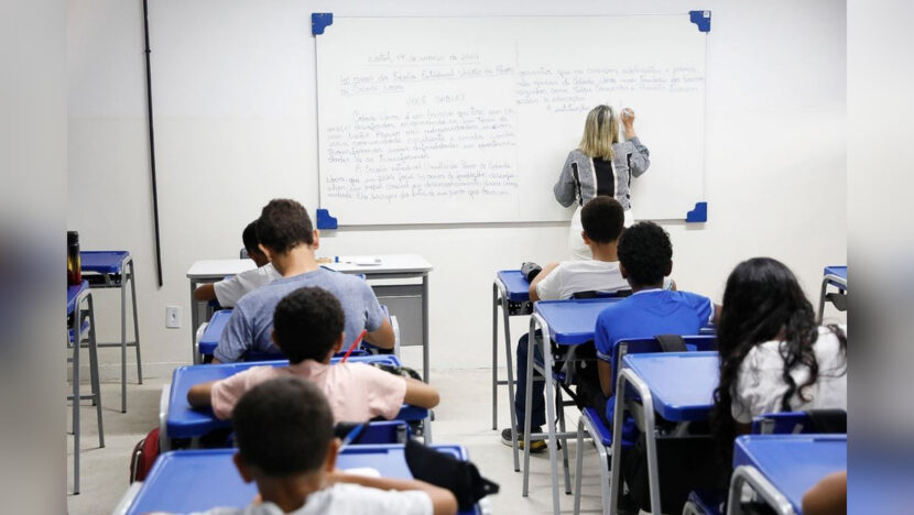Professora escreve no quadro diante de estudantes em uma sala de aula