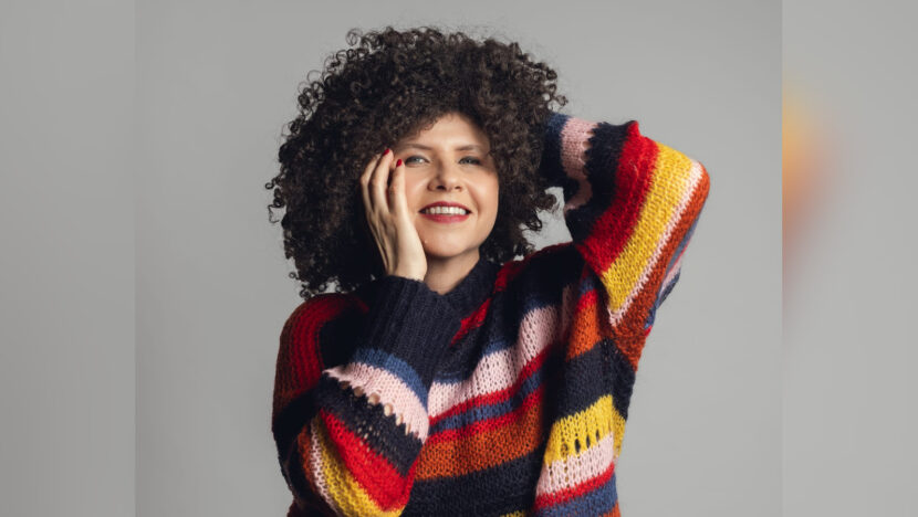 Roberta Campos, de cabelos cacheados e blusa colorida, posa com uma mão no rosto