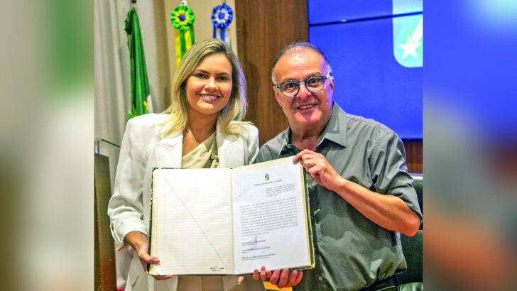 Joanna Guerra e Paulinho Freire seguram um documento e aparecem sorrindo em frente às bandeiras