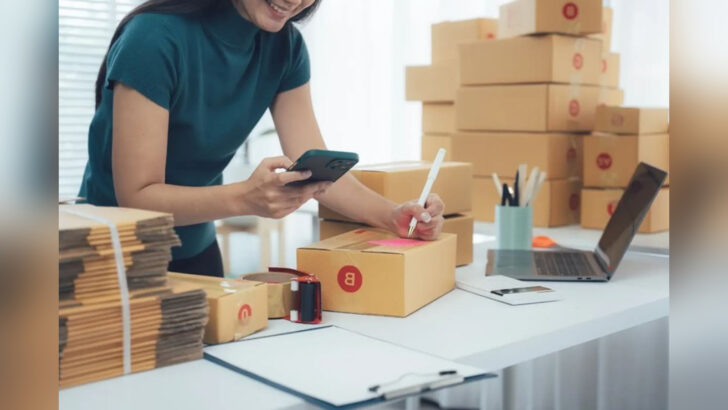 Mulher usa celular e escreve sobre uma caixa de papelão em mesa com notebook e embalagens