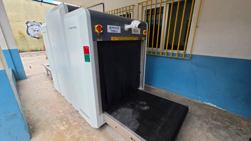 Equipamento de inspeção por raio-x instalado em unidade prisional, diante de parede azul e janela com grade amarela