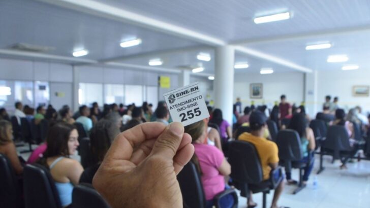 Pessoa segura senha de atendimento do Sine-RN em sala com trabalhadores