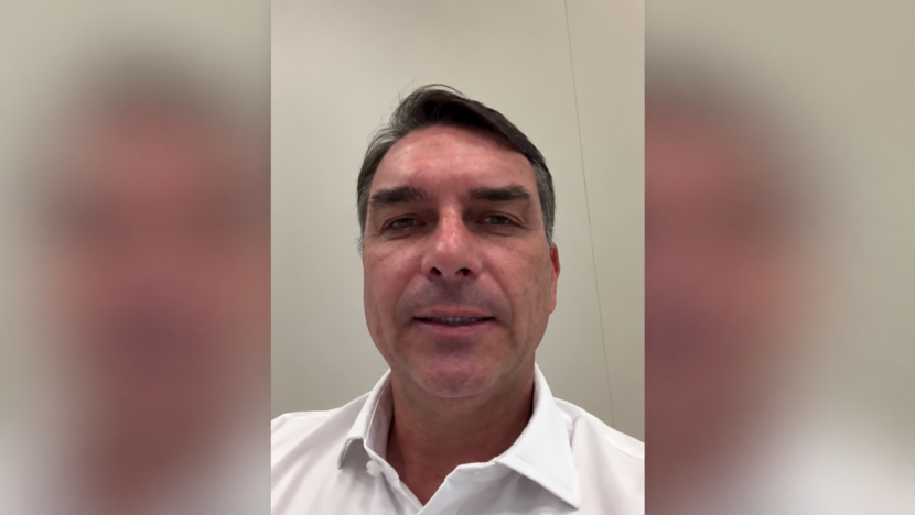 Flávio Bolsonaro aparece de camisa branca em imagem de vídeo, centralizada em formato horizontal com laterais desfocadas