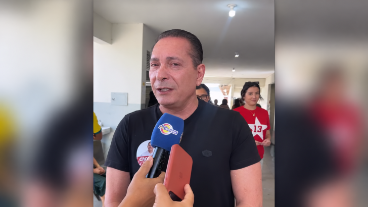 Ezequiel Ferreira, de camiseta preta, fala a um microfone durante entrevista, com pessoas ao fundo e laterais desfocadas
