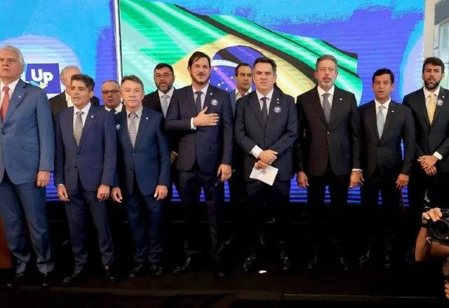 Grupo de dirigentes políticos de terno posa para fotografia diante de um painel com a bandeira do Brasil e símbolos partidários, durante evento em ambiente fechado