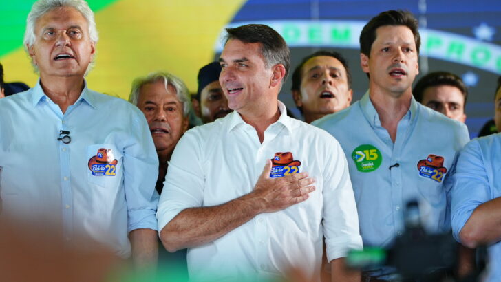 Flávio Bolsonaro segura microfone ao lado de Ronaldo Caiado e outros participantes em evento em Goiânia.