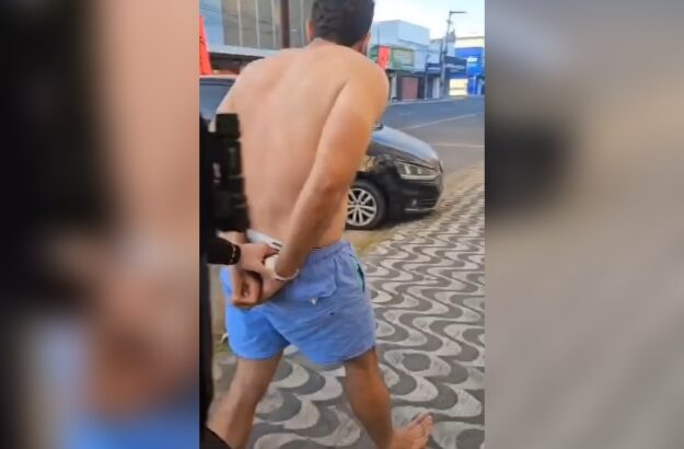 Homem sem camisa, usando bermuda azul e descalço, aparece de costas, com as mãos algemadas, sendo acompanhado por outra pessoa em uma calçada