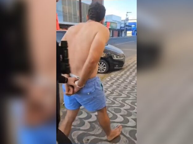 Homem sem camisa, usando bermuda azul e descalço, aparece de costas, com as mãos algemadas, sendo acompanhado por outra pessoa em uma calçada