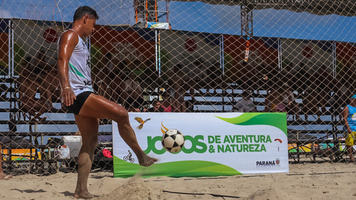 Jogador de futevôlei toca a bola com o pé em quadra de areia, junto à rede