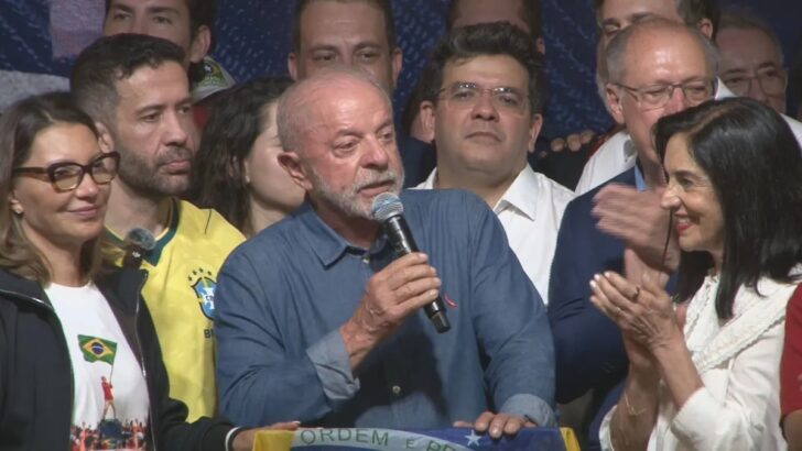 Lula fala ao microfone, segurando uma bandeira do Brasil, ao lado de Janja, Geraldo Alckmin e aliados após a reeleição