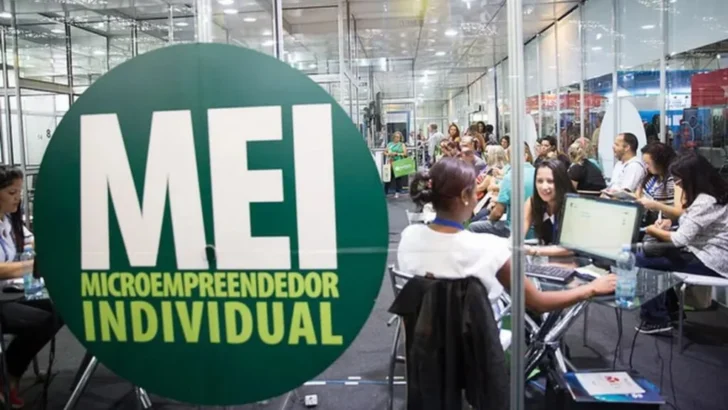 Placa circular verde com a inscrição “MEI – Microempreendedor Individual” em primeiro plano, enquanto pessoas recebem atendimento em computadores durante um evento ao fundo
