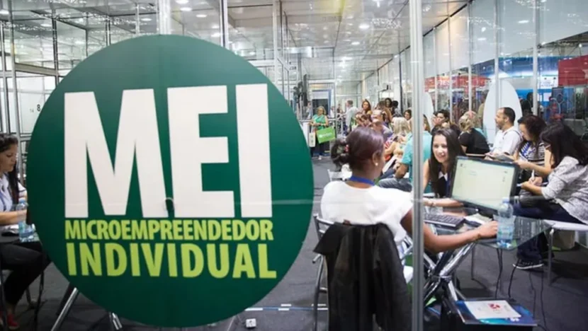 Placa circular verde com a inscrição “MEI – Microempreendedor Individual” em primeiro plano, enquanto pessoas recebem atendimento em computadores durante um evento ao fundo