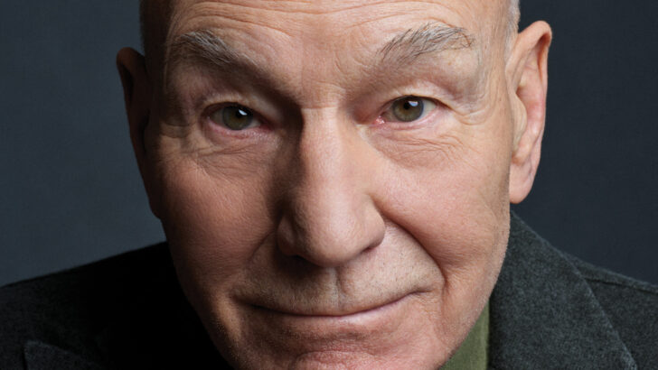 Patrick Stewart, de paletó escuro e camisa verde, olha para a câmera com as mãos unidas sob o queixo