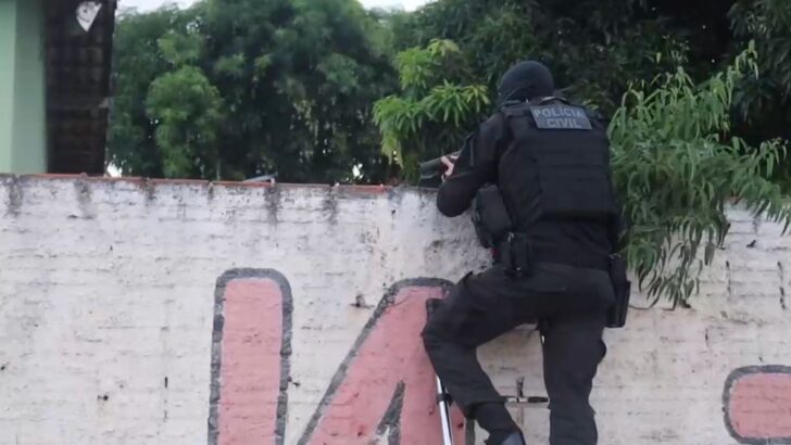 Agente da Polícia Civil, vestido com uniforme tático preto e colete de proteção, aparece apoiado em um muro durante uma ação policial. Árvores e vegetação aparecem ao fundo
