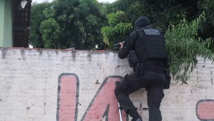 Agente da Polícia Civil, vestido com uniforme tático preto e colete de proteção, aparece apoiado em um muro durante uma ação policial. Árvores e vegetação aparecem ao fundo