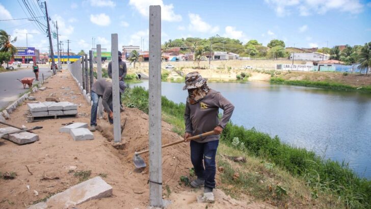Trabalhadores realizam escavações e instalam postes de concreto ao longo da margem de uma lagoa, com materiais de construção espalhados pelo terreno
