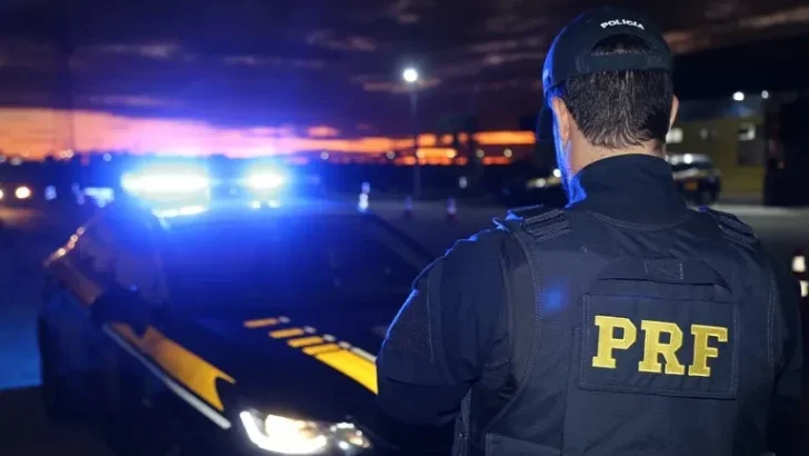 Agente da Polícia Rodoviária Federal (PRF), visto de costas e usando uniforme preto, aparece ao lado de uma viatura com luzes de emergência acionadas, durante a noite.